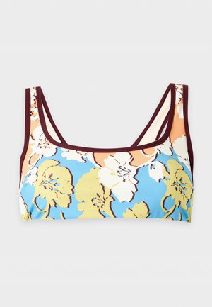 Ung kvinne står barbent iført en tofarget blomstret bikini i brune, blå, gule og hvite farger på ensfarget bakgrunn.