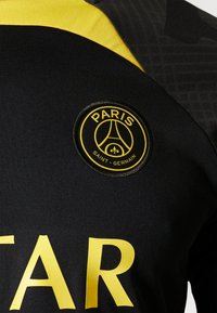 Camiseta deportiva negra con un logo circular amarillo que dice "PARIS" y "SAINT-GERMAIN." Detalles amarillos y letras grandes mejoran la visibilidad.
