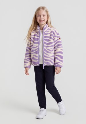 Jeune fille debout et souriante, portant une veste polaire à rayures zèbre violettes et blanches, un pantalon foncé et des baskets blanches.