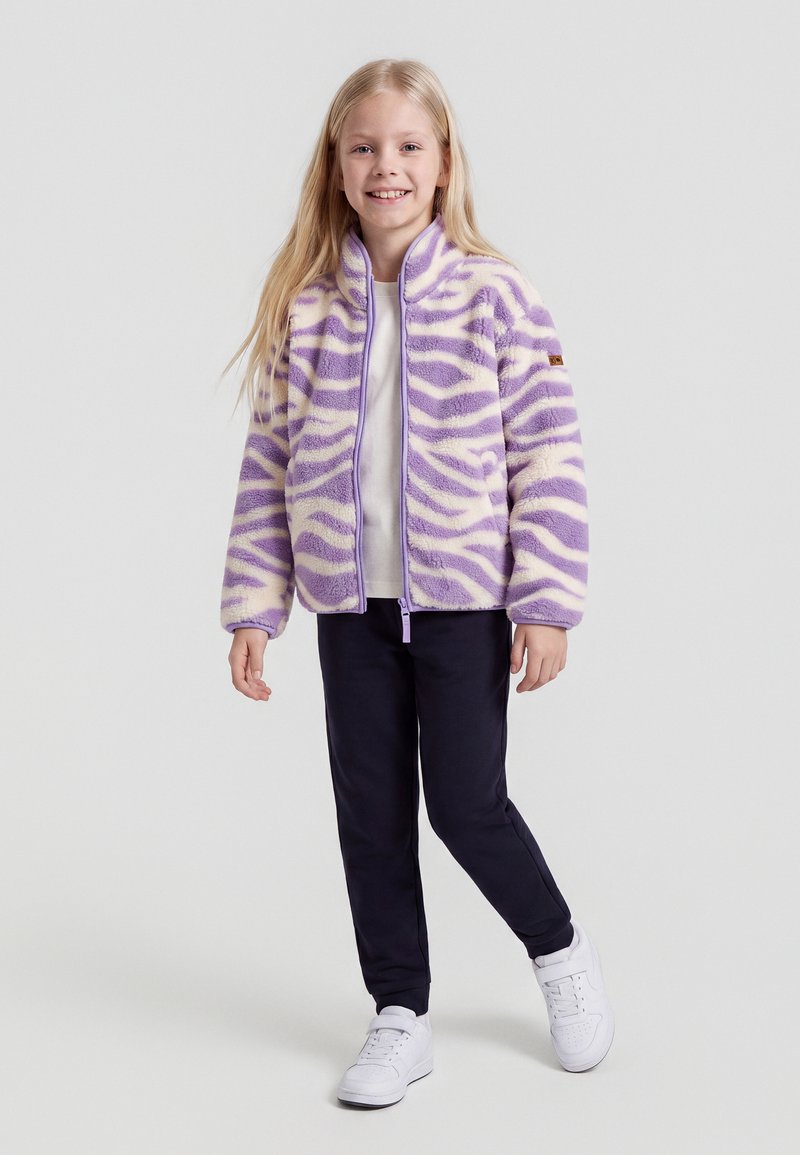 Jeune fille debout et souriante, portant une veste polaire à rayures zèbre violettes et blanches, un pantalon foncé et des baskets blanches.