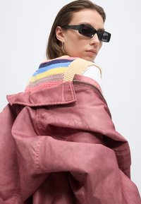 Femme portant des lunettes de soleil rectangulaires noires, un gilet en tricot à rayures multicolores, et une veste rose poudré oversized posée sur les épaules.