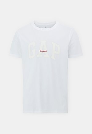 Valkoinen puuvillapaita, jossa on suuri, tekstuuri "GAP" -logo vaaleassa kermassa ja pienempi punainen "Original" teksti alla. Lyhyet hihat ja pyöreä kaula-aukko.