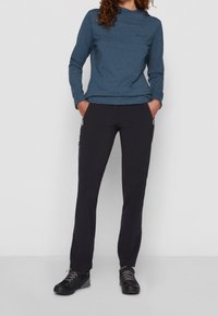 Camiseta de manga larga verde azulado con un patrón sutil, combinada con pantalones negros ajustados. Ambos hechos de materiales flexibles, con bolsillos con cremallera.