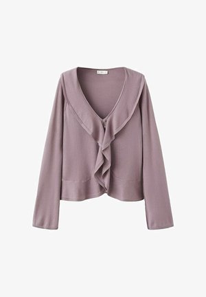 Mladá žena v fialkastej blúzke s volánmi, bielej denimovej sukni a čiernych balerínach stojaca pred jednoliatym bielym pozadím.