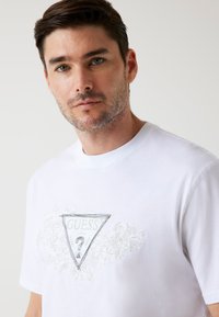 Hvid bomulds t-shirt med et trekantet "GUESS?" logo med blomsterudsmykninger i sølvbroderi. Rund halsudskæring og korte ærmer.
