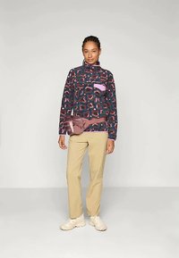 Žena stojaca v vzorovanom tmavom pulóvri s ružovým detailom na vrecku, béžových nohaviciach, bielych teniskách a mauve pásovej taške.