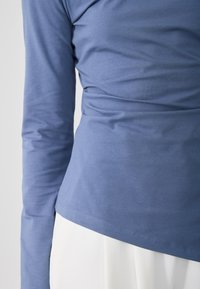 Dugi rukav u svijetloplavoj boji, izrađen od mekog materijala. Ima prilagođen dizajn s naborima na struku i ravnim rubom.
