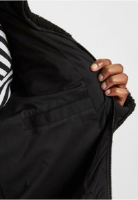 Das Innere der schwarzen Jacke besteht aus einem glatten, strukturierten Stoff mit einem Mesh-Muster, der einen Reißverschluss und eine Seitentasche zeigt.