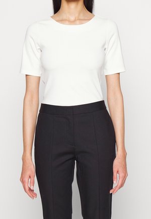 Kobieta ubrana w biały dopasowany top z krótkim rękawem włożony w czarne eleganckie spodnie z wysokim stanem, na jednolitym tle.