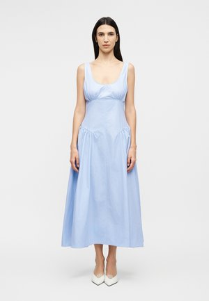 Γυναίκα με μακριά σκούρα μαλλιά στέκεται μπροστά, φοράει αμάνικο φωτεινό γαλάζιο μεσαίο φόρεμα και άσπρες μυτερές γόβες, σε απλό φόντο.