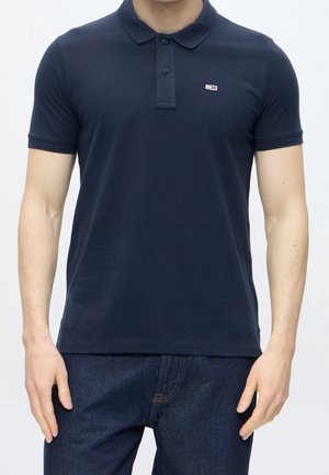 Mand iført en marineblå poloshirt med korte ærmer, krave og knaplukning, kombineret med mørkeblå jeans.