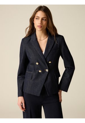 Donna che indossa un blazer doppiopetto in denim scuro con bottoni dorati, abbinato a pantaloni scuri, in piedi davanti a uno sfondo beige uniforme.