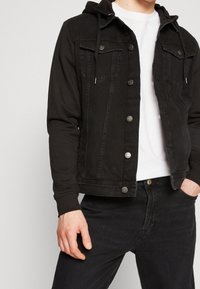 Svart denimjacka med huva, med metallknappar, två bröstfickor och kontrasterande ärmtyg. Bärs över en vit t-shirt.