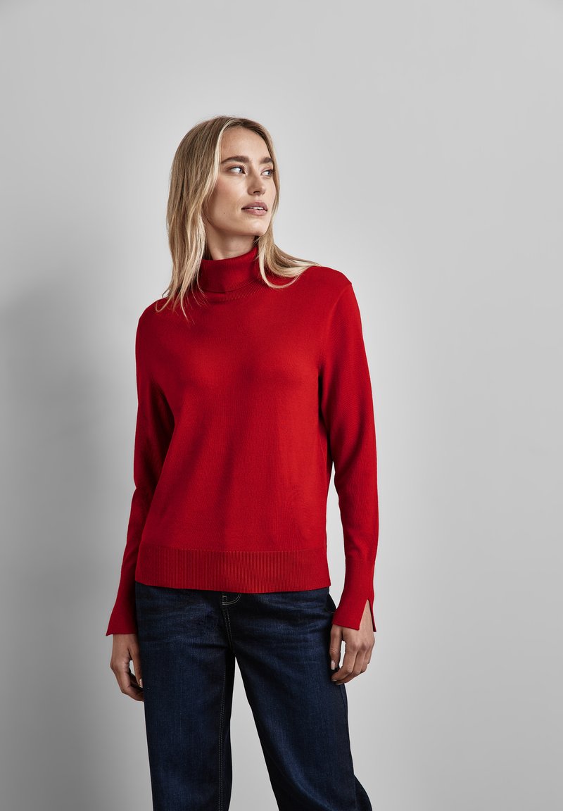 Roter Rollkragenpullover aus weichem Stoff mit gerippten Bündchen und Saum. Kombiniert mit dunkelblauen Jeans, die einen lockeren Schnitt und seitliche Schlitze aufweisen.