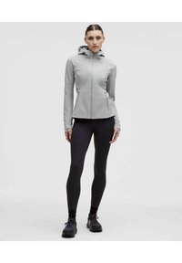 Chaqueta gris claro con cremallera, capucha, mangas largas y dos bolsillos delanteros. Combinada con leggings negros y zapatillas deportivas oscuras.