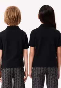 Två barn står med ryggen mot kameran iförda svarta kortärmade polo-tröjor och mönstrade grå byxor mot en enfärgad bakgrund.