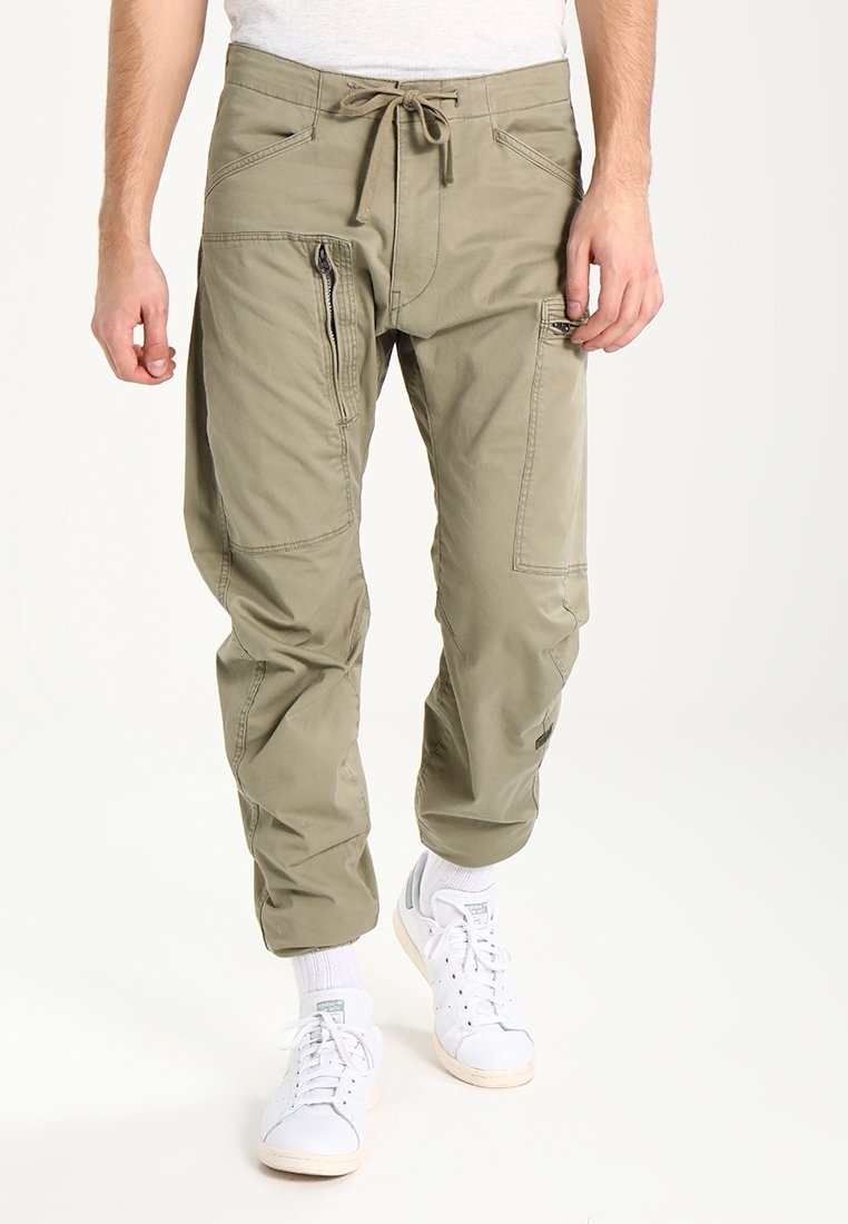 Uomo che indossa pantaloni cargo beige con tasche con zip e sneakers bianche, in piedi di fronte a uno sfondo uniforme.