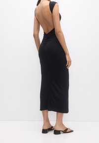 Robe noire moulante avec un dos décolleté, des détails froncés à la taille et des manches courtes. Portée avec des sandales à talons ouvertes.