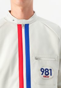 Nærbilde av en person iført en lys grå jakke med røde og blå striper og en sirkulær lapp med "981 REPLAY" på lommen.