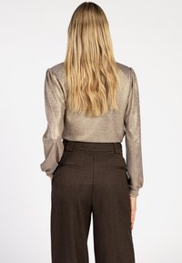Blonde vrouw met lang haar, gekleed in een gouden top met lange mouwen en hoge taille donkere brown wijde broek, staande met de rug naar de kijker gericht.