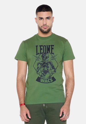 Mann mit kurzen dunklen Haaren und Bart, der ein grünes T-Shirt mit schwarzer Grafik eines Skelett-Boxers und dem Text "LEONE 1947 WACS." trägt.