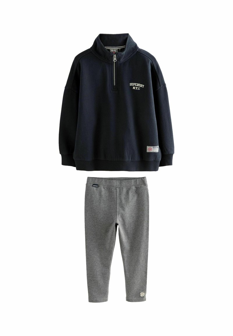Námornícky modrá mikina s krátkym zipsom s textom "Superdry NYC" v kombinácii so sivými priliehavými legínami, obidva voľnočasové kúsky.