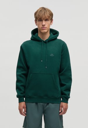 Joven con cabello castaño claro que lleva una sudadera con capucha verde oscuro con bolsillo delantero y pantalones cortos grises, de pie frente a un fondo liso.