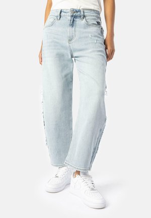 Jeans azzurri chiari a vita alta con vestibilità rilassata, dettagli strappati e orli sfrangiati, abbinati a sneakers bianche.