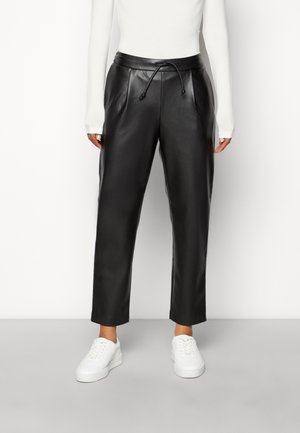 Pantalons de jogging en cuir noir avec une taille à cordon, un devant plissé et des jambes fuselées, associés à des baskets blanches sur un fond gris clair.