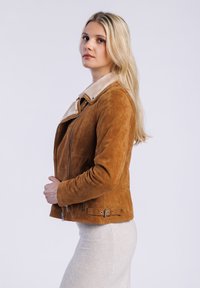Eine Frau mit langen blonden Haaren trägt eine braune Wildlederjacke mit Kunstfellkragen und posiert seitlich vor einem schlichten weißen Hintergrund.