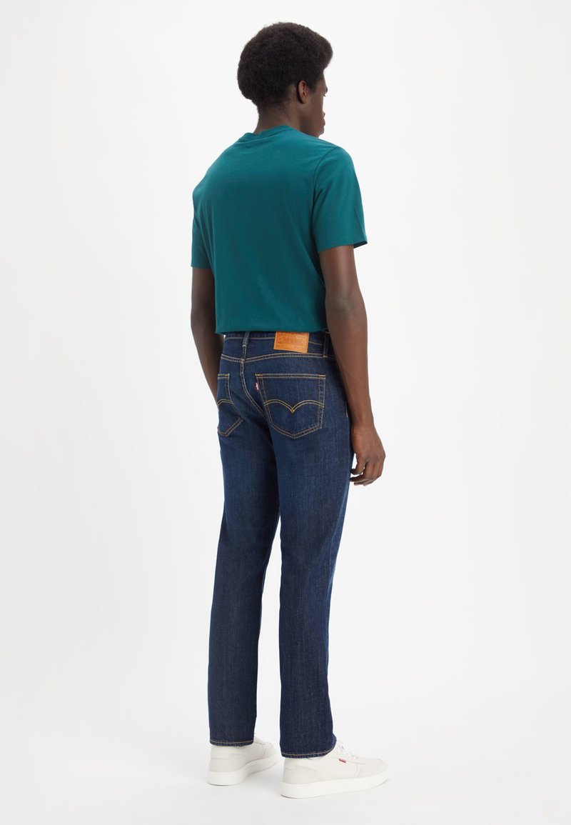 Camiseta de manga corta en couleur verde azulado combinada con jeans rectos de color azul oscuro, que presentan una etiqueta de cuero en la parte posterior de la cintura y zapatillas blancas.