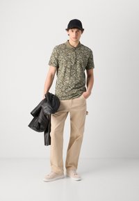 Grünes Polohemd mit floralem Muster, beigen Hosen, schwarze Lederjacke über dem Arm, schwarze Mütze, cremefarbene Sneaker, lässiges Outfit.