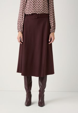 Femme portant une jupe marron longueur genoux avec une ceinture assortie, un chemisier à manches longues à motifs et des bottes en cuir marron montant jusqu'aux genoux, debout bien droite.