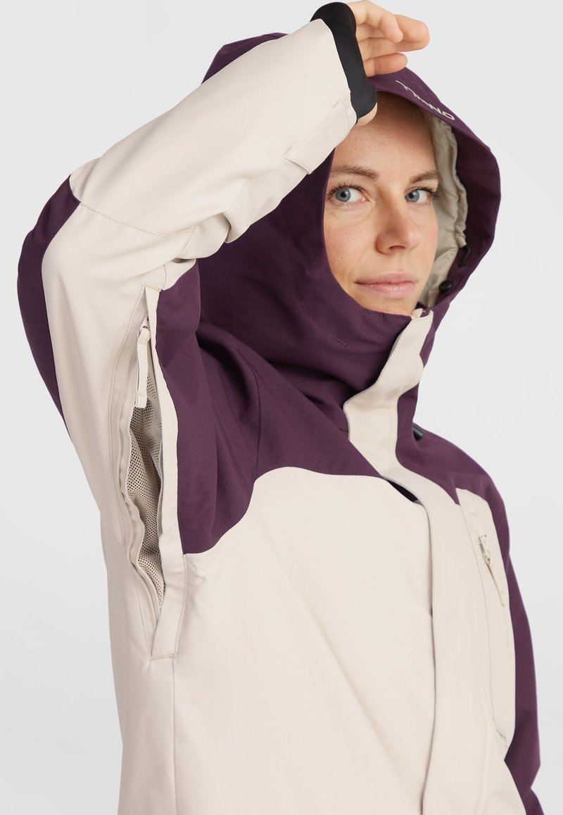 Jacke mit einem creme- und lila-farbenen Blockdesign, verstellbare Kapuze, sichtbarem Reißverschluss und Netzbelüftungsdetails für Atmungsaktivität.