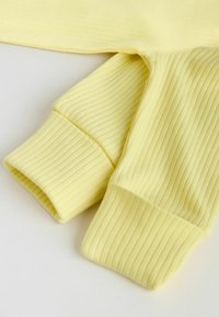 Material moale galben cu nervuri, prezentând o prim-planare a mânecilor lungi pliate cu manșete late pe o suprafață albă.
