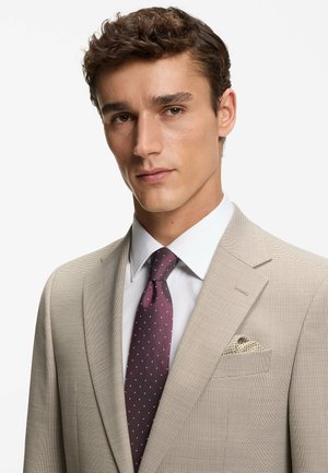 Giacca di completo beige testurizzata con revers a lancia, camicia bianca e cravatta viola scuro con pois bianchi. Fazzoletto da taschino con motivo a tessuto intrecciato.