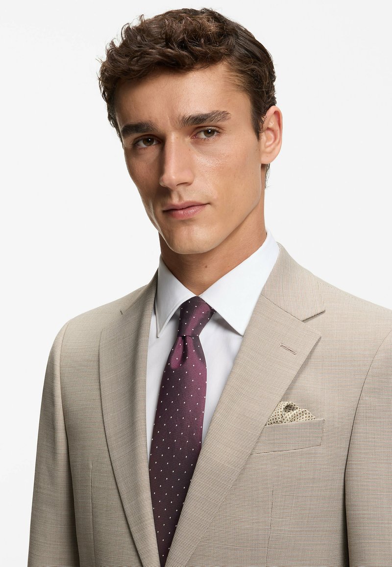 Giacca di completo beige testurizzata con revers a lancia, camicia bianca e cravatta viola scuro con pois bianchi. Fazzoletto da taschino con motivo a tessuto intrecciato.