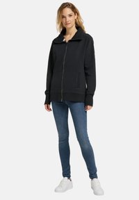 Schwarze Zip-up-Sweatjacke mit hohem Kragen, gerippten Bündchen und zwei frontalen Taschen, kombiniert mit blauen skinny Jeans und weißen Sneakers.