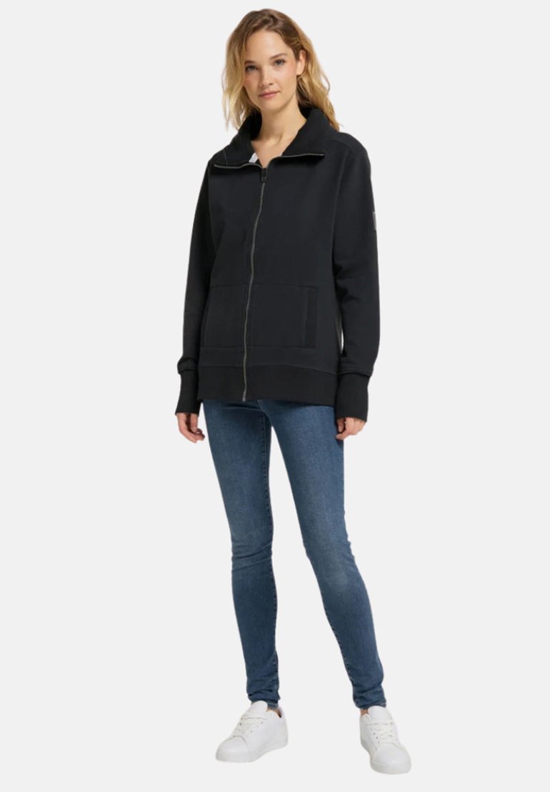 Schwarze Zip-up-Sweatjacke mit hohem Kragen, gerippten Bündchen und zwei frontalen Taschen, kombiniert mit blauen skinny Jeans und weißen Sneakers.