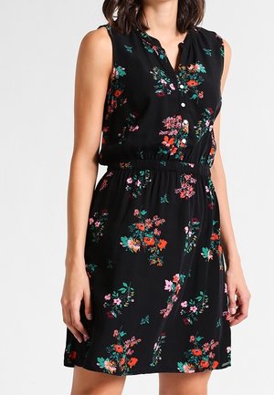 Ærmeløs sort kjole med farverigt blomsterprint, knaplukning foran og elastisk taljebånd, båret af en person mod en ensfarvet baggrund.
