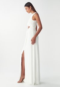Vestido blanco sin mangas con una alta abertura, textura drapeada y una sección media con corte. Llevado con sandalias nude de tiras.