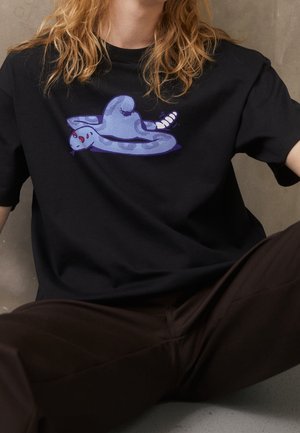 Personne portant un t-shirt noir avec un serpent de dessin animé violet aux yeux rouges allongé à plat, portant un pantalon foncé.