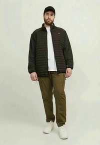 Homem com barba a usar um boné preto, um casaco acolchoado preto, uma camisa branca, umas calças cargo verde-oliva e uns ténis brancos, de pé contra um fundo claro.