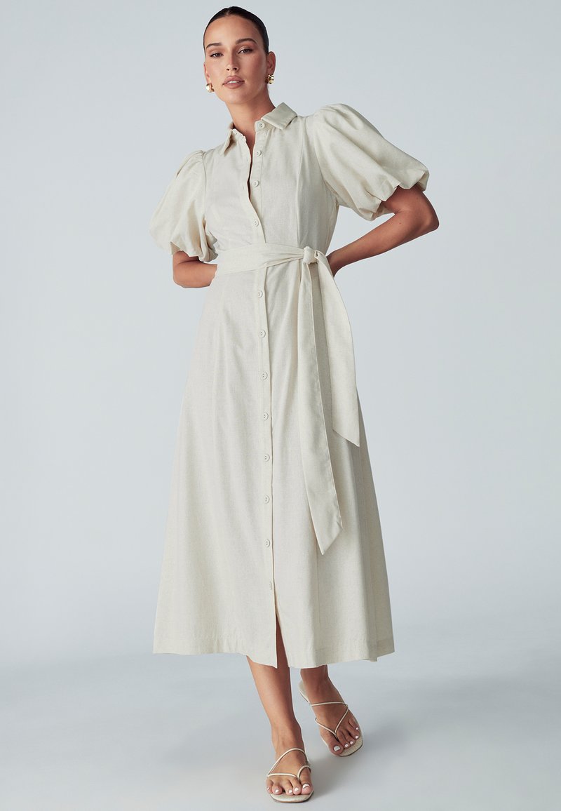 Šviesiai beigės spalvos, midi ilgio marškininė su pūstomis rankovėmis, priekiniais mygtukais ir suderintu juosmens diržu. Pora su kaspinėmis basutėmis.