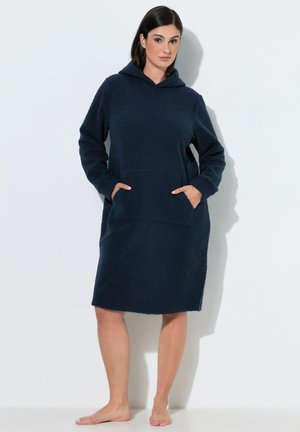 Femme portant une robe polaire à capuche bleu foncé à manches longues avec poches avant, debout pieds nus sur un fond clair uni.