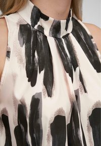 Blouse sans manches en tissu léger et lisse, avec un col haut. Elle est de couleur beige avec des motifs audacieux de coups de pinceau noirs.