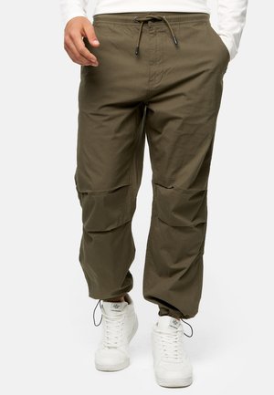 Pantalones cargo verde oliva con cintura elástica, cierre de cordón, dos bolsillos laterales y detalles de parches en las rodillas. Combinados con zapatillas blancas.