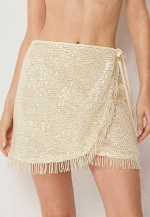 Beige Wickel-Minirock mit Pailletten und Fransen, an der Taille gebunden, getragen von einer stehenden Person.