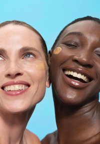 Twee individuen met een gladde huid, de een met een lichte foundation en de ander met een donkerdere tint, lachend tegen een blauwe achtergrond.