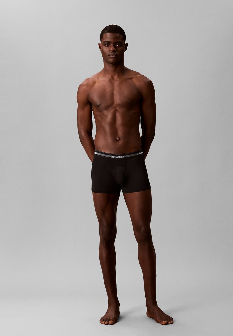 Homme musclé debout pieds nus sur fond gris, portant un boxer noir Calvin Klein, les bras derrière le dos, face à l’avant.
